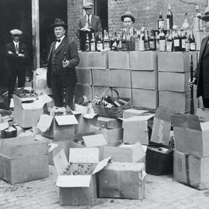May include: A black and white photo of a group of men standing in front of a pile of boxes filled with bottles. The boxes are stacked in a wall behind the men. The men are wearing suits and hats. The bottles are in clear glass and are arranged in rows. The photo is likely from the 1920s and depicts a scene of illegal alcohol production or distribution during the Prohibition era.