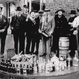 May include: A group of people stand in front of a brick building. They are holding a variety of glass bottles and jugs. There is a wooden barrel in the center of the image. The bottles and jugs are filled with a clear liquid. There is a bucket and a box of wooden sticks in the foreground.