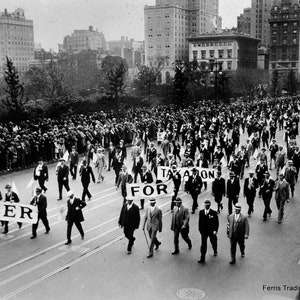 May include: A black and white photo of a large crowd of people marching down a city street. The people are holding signs that say 