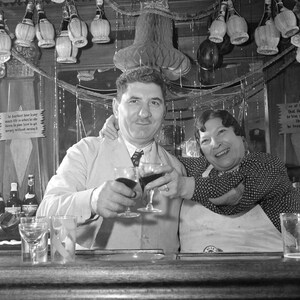 May include: Two people are sitting at a bar counter. The person on the left is wearing a white shirt and a tie. The person on the right is wearing a dark dress and a white apron. Both people are holding glasses of wine. The bar is decorated with string lights and there are bottles of wine on the counter. There is a sign on the wall that reads 