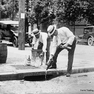 May include: Three men in white shirts and hats are pouring bottles of liquor into a sewer drain. The bottles are arranged on the sidewalk in front of the drain. There are two barrels in the background. The image is in black and white. Ferris Trading Company is printed in the lower right corner of the image.