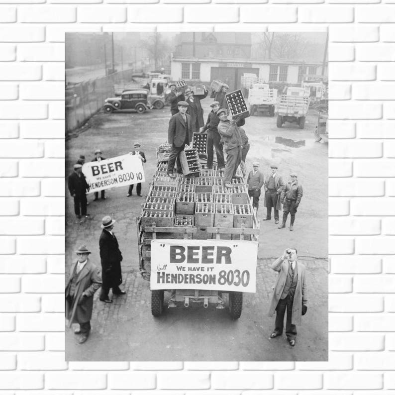A vintage black and white photograph of men in flat caps and coats loading crates of beer onto a truck in Cleveland, Ohio. A large banner on the back of the truck reads "BEER" Call WE HAVE IT HENDERSON 8030.