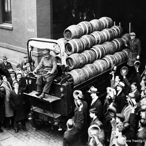 May include: A black and white photo of a group of people gathered around a truck loaded with wooden barrels. The truck is parked in front of a brick building with the number 1609 visible. The people are all wearing hats and are smiling and waving. The truck is labeled 