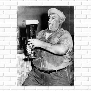 May include: A man in a light-colored shirt and dark pants holds a tall glass of beer with a thick head of foam.