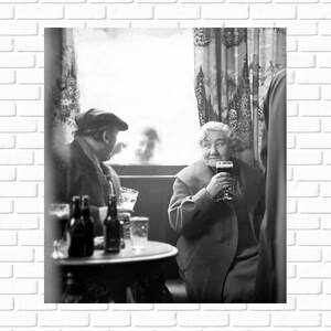 May include: A black and white photo of two people sitting at a table in a pub. The person on the left is wearing a hat and is looking down at a glass. The person on the right is holding a glass of beer and looking at the camera.