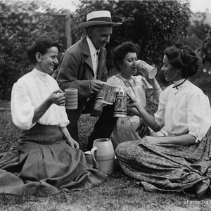 May include: Four people are sitting on the grass, drinking from mugs. They are all wearing light-colored clothing. The person on the left is wearing a white blouse and a dark skirt. The person in the middle is wearing a light-colored dress. The person on the right is wearing a white blouse and a dark skirt. The person in the back is wearing a light-colored shirt and dark pants. The people are all smiling and looking at the camera. The grass is green and there are trees in the background. The text 