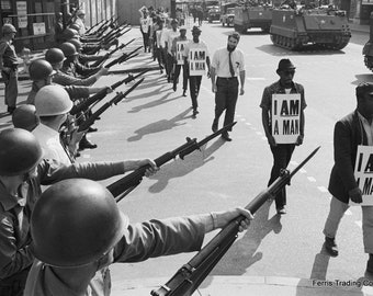 May include: Black and white photo of a protest. A line of men in military uniforms hold rifles and face a group of men holding signs that read "I am a man".