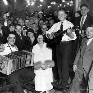 May include: A black and white photo of a group of people gathered together. A person plays an accordion and another person plays a guitar. The people are smiling and appear to be enjoying themselves. The photo is taken in a dimly lit room with a sign that reads 