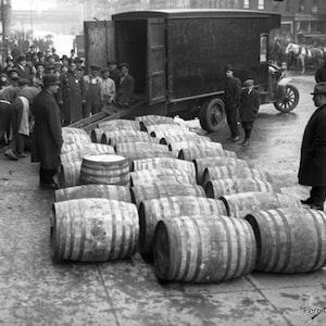 May include: A black and white photo of a street scene with a truck unloading barrels. The truck has the words 