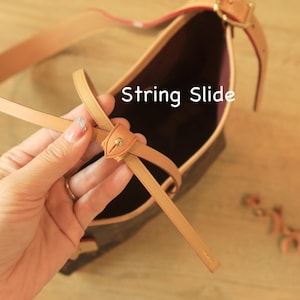 Puede incluir: Bolso de mano de cuero marrón claro con una correa a juego y herrajes dorados. El bolso presenta un detalle de deslizamiento de cuerda. El interior del bolso es de color morado oscuro.