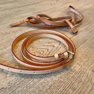 Op de afbeelding: Een opgerolde, lichtbruine leren riem met een goudkleurige gesp. De riem heeft een dun ontwerp met een contrasterende donkerbruine rand en witte stiksels. De riem ligt op een houten oppervlak.