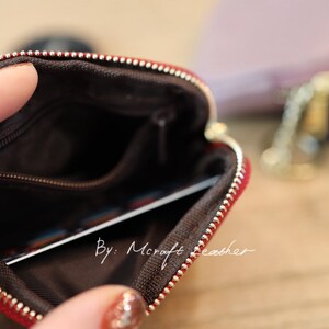 Puede incluir: Una bolsa de cuero rojo con cremallera y forro marr&oacute;n. La bolsa est&aacute; abierta y se puede ver una tarjeta en el interior. El texto "By: Mcraft Leather" est&aacute; impreso en la bolsa.