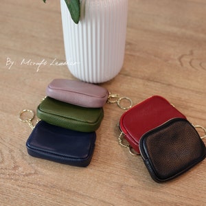 Puede incluir: Cinco peque&ntilde;as carteras de monedas de cuero con llaveros dorados. Las carteras est&aacute;n apiladas en forma de pir&aacute;mide, con la cartera superior de color morado, la siguiente verde, la siguiente azul marino, la siguiente roja y la cartera inferior negra.
