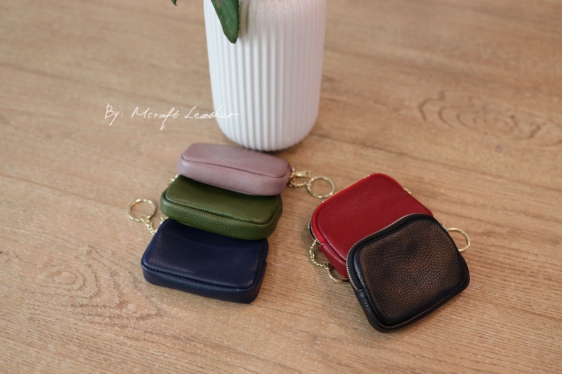 Puede incluir: Cinco peque&ntilde;as carteras de monedas de cuero con llaveros dorados. Las carteras est&aacute;n apiladas en forma de pir&aacute;mide, con la cartera superior de color morado, la siguiente verde, la siguiente azul marino, la siguiente roja y la cartera inferior negra.