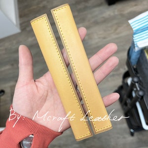 May include: Two yellow leather straps with decorative stitching. The straps are lined up side by side and have a slight curve to them. The text "By: Mcroft Leather" is visible in the image.
