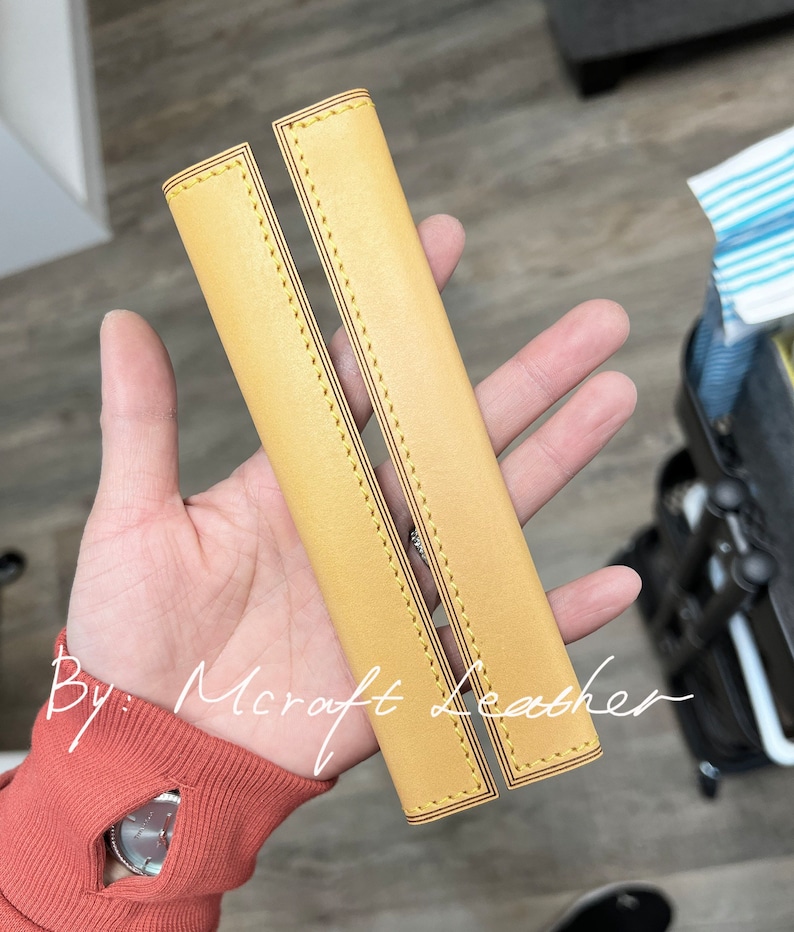 May include: Two yellow leather straps with decorative stitching. The straps are lined up side by side and have a slight curve to them. The text "By: Mcroft Leather" is visible in the image.