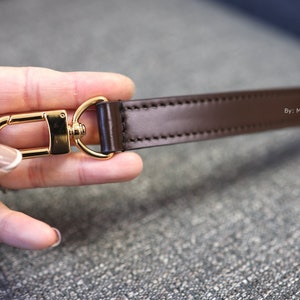 May include: A close-up of a brown leather strap with gold-toned hardware. The strap is stitched and has the text "By: Mcraft Leather" printed on it.