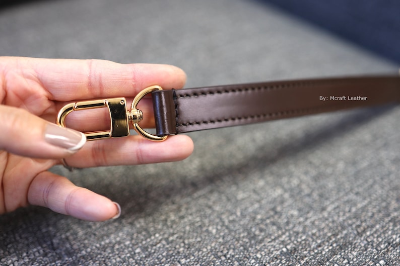 May include: A close-up of a brown leather strap with gold-toned hardware. The strap is stitched and has the text "By: Mcraft Leather" printed on it.