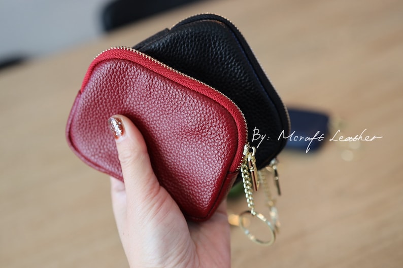 Puede incluir: Dos peque&ntilde;as carteras con cremallera, una roja y otra negra, con una cadena dorada y un llavero. Las carteras son de cuero y tienen una superficie texturizada. Por: Mcraft Leather.