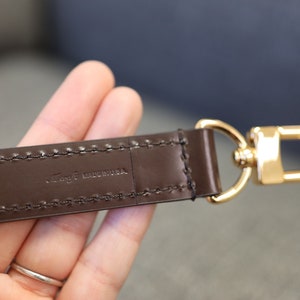 May include: A brown leather strap with a gold metal hook. The strap has stitching along the edges and the text "Togo Made in USA" is embossed on the strap.