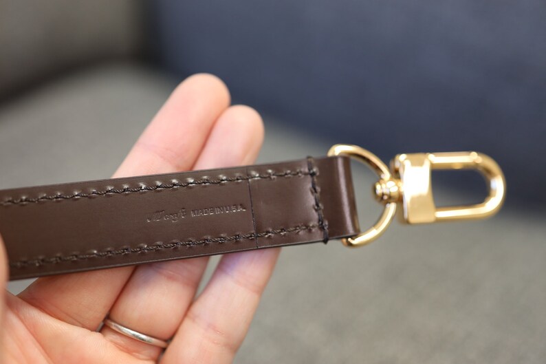 May include: A brown leather strap with a gold metal hook. The strap has stitching along the edges and the text "Togo Made in USA" is embossed on the strap.