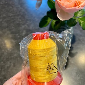 May include: A spool of bright yellow thread wrapped in clear plastic packaging. The spool has a red top and bottom and is labeled with a recycling symbol.