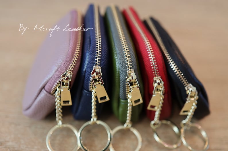 Puede incluir: Cinco peque&ntilde;as bolsas con cremallera de color dorado y llaveros. Las bolsas son de color rosa, azul, verde, rojo y negro. El texto "By: Mcraft Leather" es visible en la bolsa superior.