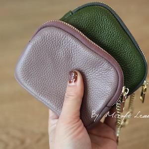 Puede incluir: Dos peque&ntilde;as carteras de monedas con cremallera, una de color rosa polvoriento y la otra de color verde bosque oscuro, se sostienen en una mano. Las carteras tienen una cadena dorada unida a ellas. El texto "By: Mcraft Leather" es visible en la esquina inferior derecha de la imagen.