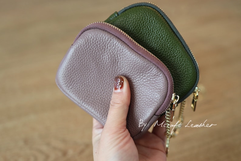 Puede incluir: Dos peque&ntilde;as carteras de monedas con cremallera, una de color rosa polvoriento y la otra de color verde bosque oscuro, se sostienen en una mano. Las carteras tienen una cadena dorada unida a ellas. El texto "By: Mcraft Leather" es visible en la esquina inferior derecha de la imagen.