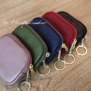 Puede incluir: Cinco peque&ntilde;as bolsas con cremallera y llaveros, en tonos de morado, verde, azul marino y rojo. Las bolsas est&aacute;n hechas de cuero y tienen un acabado texturizado. El texto "By: Mcraft Leather" es visible en la esquina superior izquierda de la imagen.