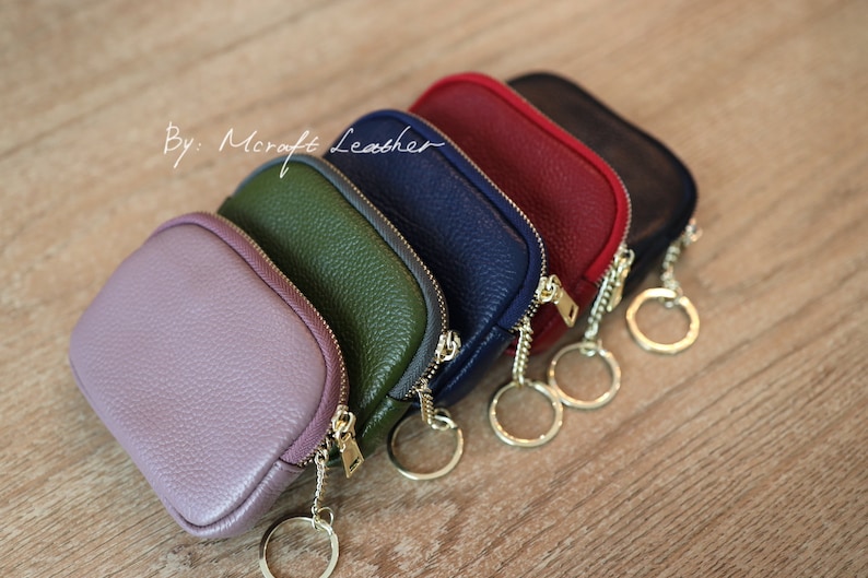 Puede incluir: Cinco peque&ntilde;as bolsas con cremallera y llaveros, en tonos de morado, verde, azul marino y rojo. Las bolsas est&aacute;n hechas de cuero y tienen un acabado texturizado. El texto "By: Mcraft Leather" es visible en la esquina superior izquierda de la imagen.