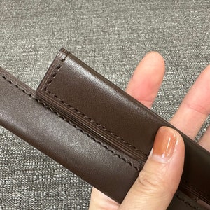 May include: Two brown leather sheaths with stitching detail. The sheaths are held in a hand.