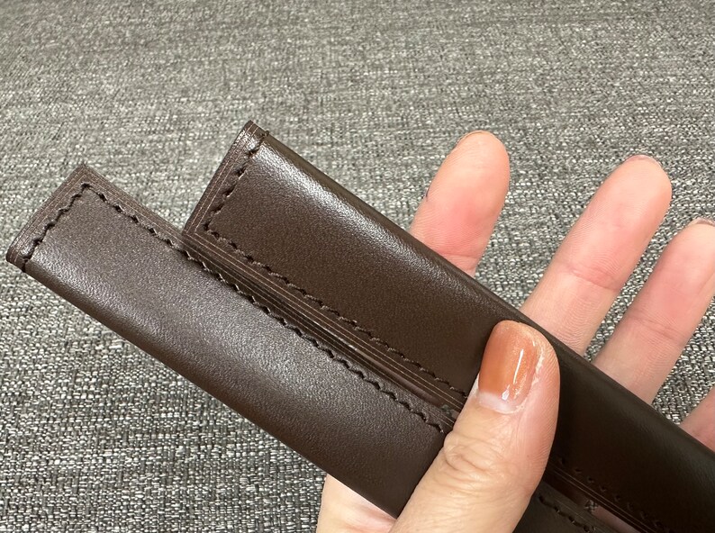 May include: Two brown leather sheaths with stitching detail. The sheaths are held in a hand.