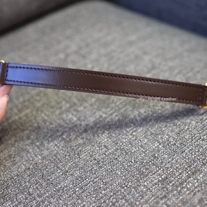 May include: A brown leather strap with gold metal hardware. The strap is stitched with a single row of stitching. The text "By: McThrift Leather" is printed on the strap.
