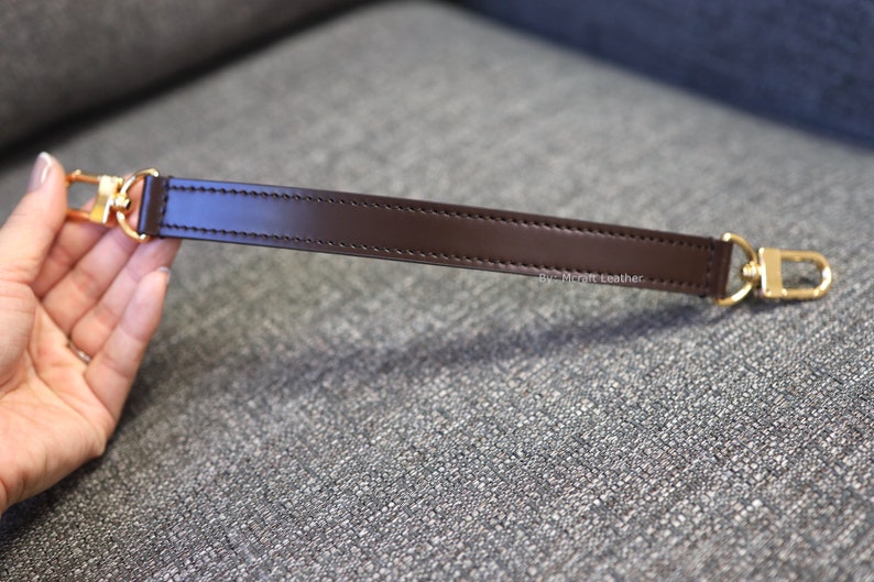 May include: A brown leather strap with gold metal hardware. The strap is stitched with a single row of stitching. The text "By: McThrift Leather" is printed on the strap.