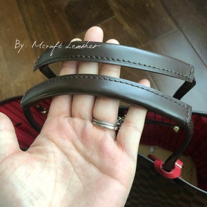 Puede incluir: Primer plano de las asas de cuero marrón de un bolso de diseño rojo y marrón. Las asas están cosidas con hilo marrón y tienen una ligera curva. El texto "By: Mcraft Leather" es visible en la esquina superior izquierda de la imagen.