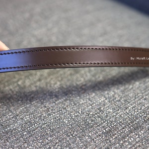 May include: A close-up of a brown leather strap with stitching and a gold metal clasp. The strap is labeled "By: Mcraft Leather".