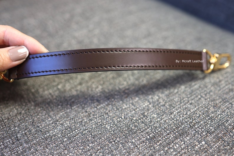 May include: A close-up of a brown leather strap with stitching and a gold metal clasp. The strap is labeled "By: Mcraft Leather".