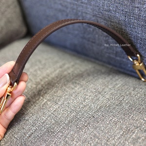 May include: A brown leather strap with gold hardware. The strap is attached to a fabric surface. The text "By: Mcraft Leather" is visible on the fabric.