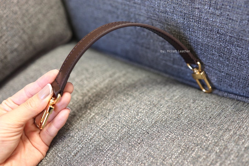 May include: A brown leather strap with gold hardware. The strap is attached to a fabric surface. The text "By: Mcraft Leather" is visible on the fabric.