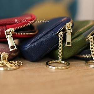Puede incluir: Un conjunto de cuatro peque&ntilde;as bolsas de cuero con cremallera, en rojo, marr&oacute;n, azul y verde, con cremalleras doradas y cadenas unidas a un llavero dorado. Las bolsas est&aacute;n etiquetadas con "By: Mcraft Leather".