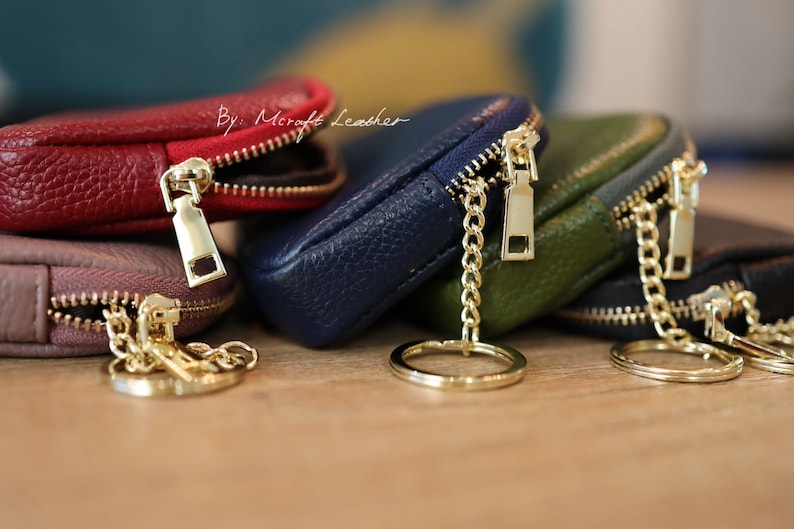 Puede incluir: Un conjunto de cuatro peque&ntilde;as bolsas de cuero con cremallera, en rojo, marr&oacute;n, azul y verde, con cremalleras doradas y cadenas unidas a un llavero dorado. Las bolsas est&aacute;n etiquetadas con "By: Mcraft Leather".