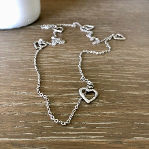 May include: A silver chain necklace with multiple heart-shaped charms. The hearts are textured and have a slightly rustic look.