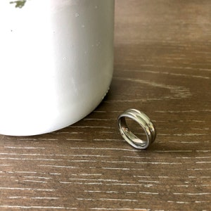 May include: A silver-toned metal ring with a smooth, polished finish and a subtle central groove running along the circumference of the band. The ring is positioned upright, showcasing its circular shape and reflective metallic surface.