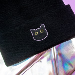 Op de afbeelding: Zwarte beanie met een roze geborduurd kattengezicht met gele ogen.