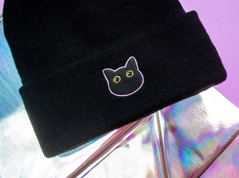 Op de afbeelding: Zwarte beanie met een roze geborduurd kattengezicht met gele ogen.