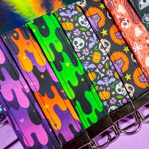May include: A set of six colorful keychains with different Halloween-themed designs. The keychains feature a variety of patterns, including slime, bats, skulls, and cats. Each keychain has a silver metal ring.