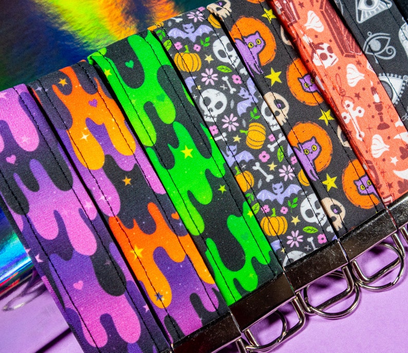 May include: A set of six colorful keychains with different Halloween-themed designs. The keychains feature a variety of patterns, including slime, bats, skulls, and cats. Each keychain has a silver metal ring.