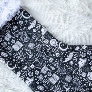 May include: A black and white Christmas stocking with a spooky, witchy design. The stocking features a variety of illustrations including crystals, potion bottles, bats, and moons.