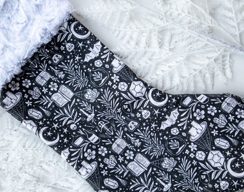 May include: A black and white Christmas stocking with a spooky, witchy design. The stocking features a variety of illustrations including crystals, potion bottles, bats, and moons.